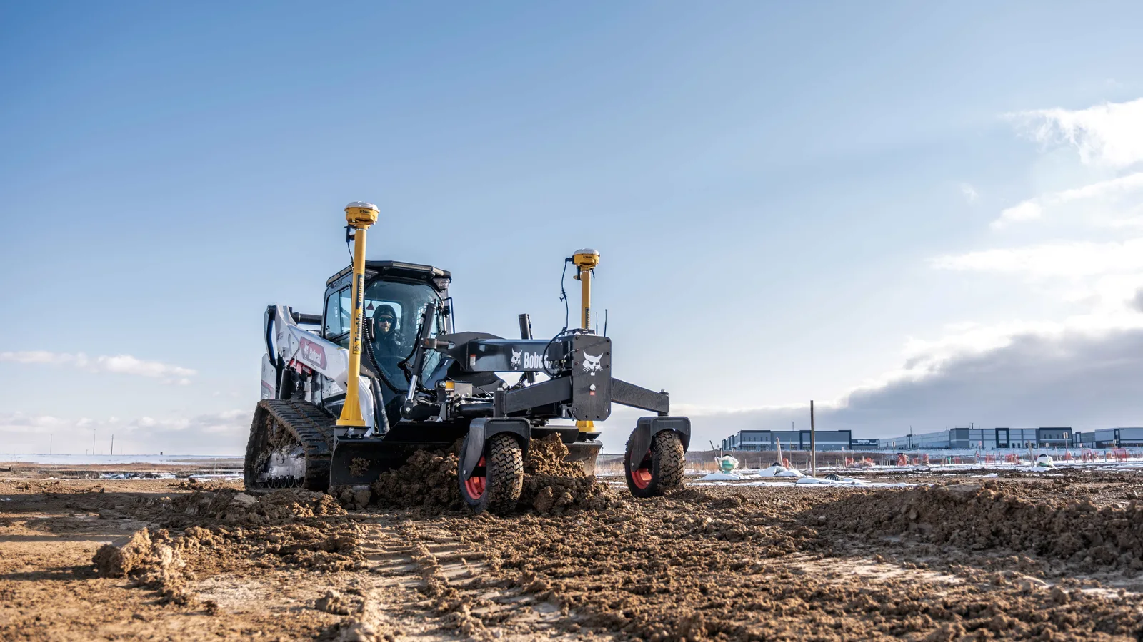 Bobcat compact track loader running a grader attachment, leveling a wide dirt pad on an open building site under a clear sky