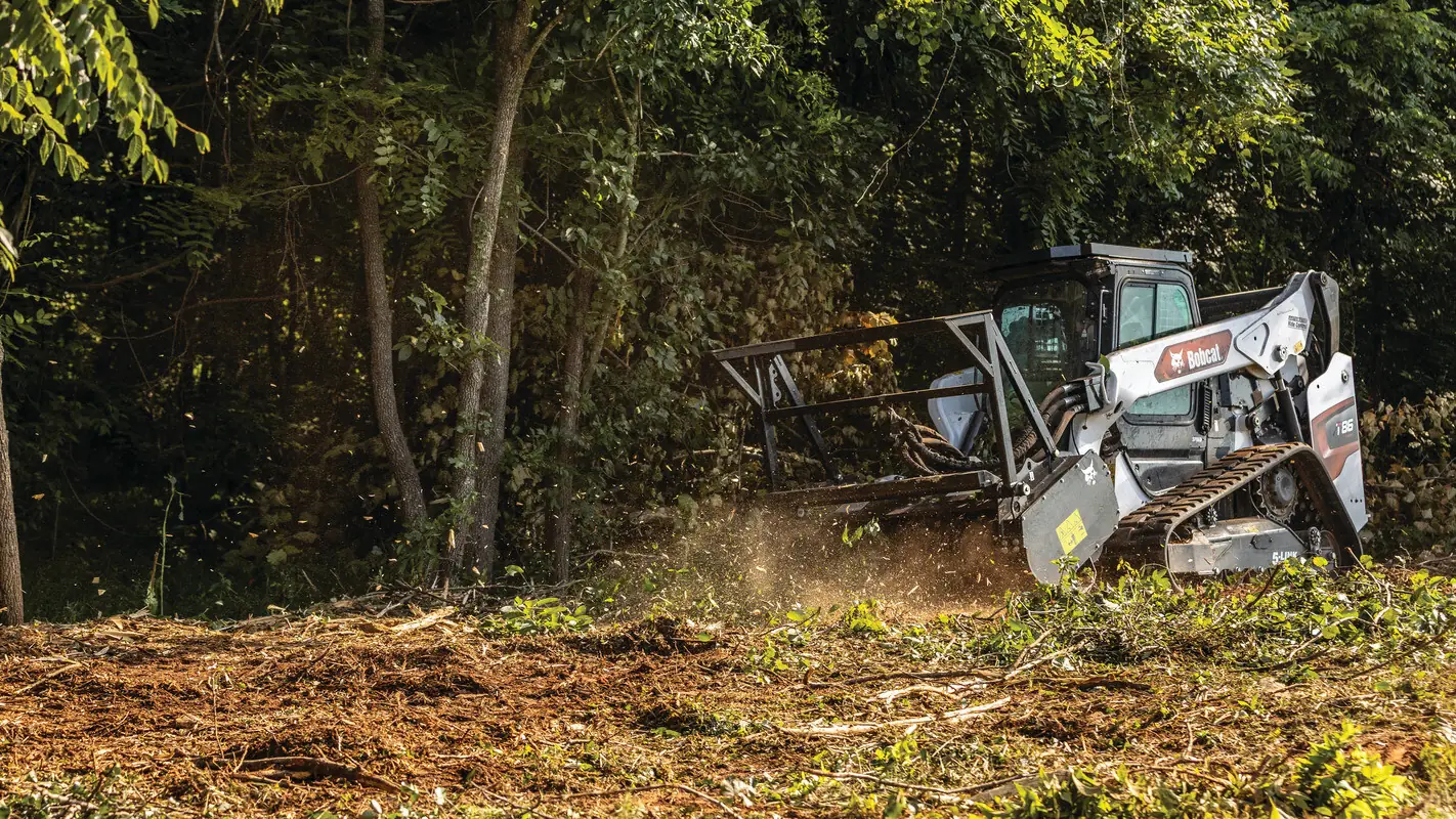 Compact track loader running a forestry mulcher head into brush at the edge of the woods, with mulch flying