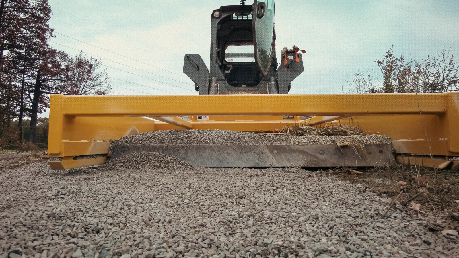 Low view along a gravel driveway as a skid steer pulls a grader blade across the surface, smoothing loose rock into an even lane