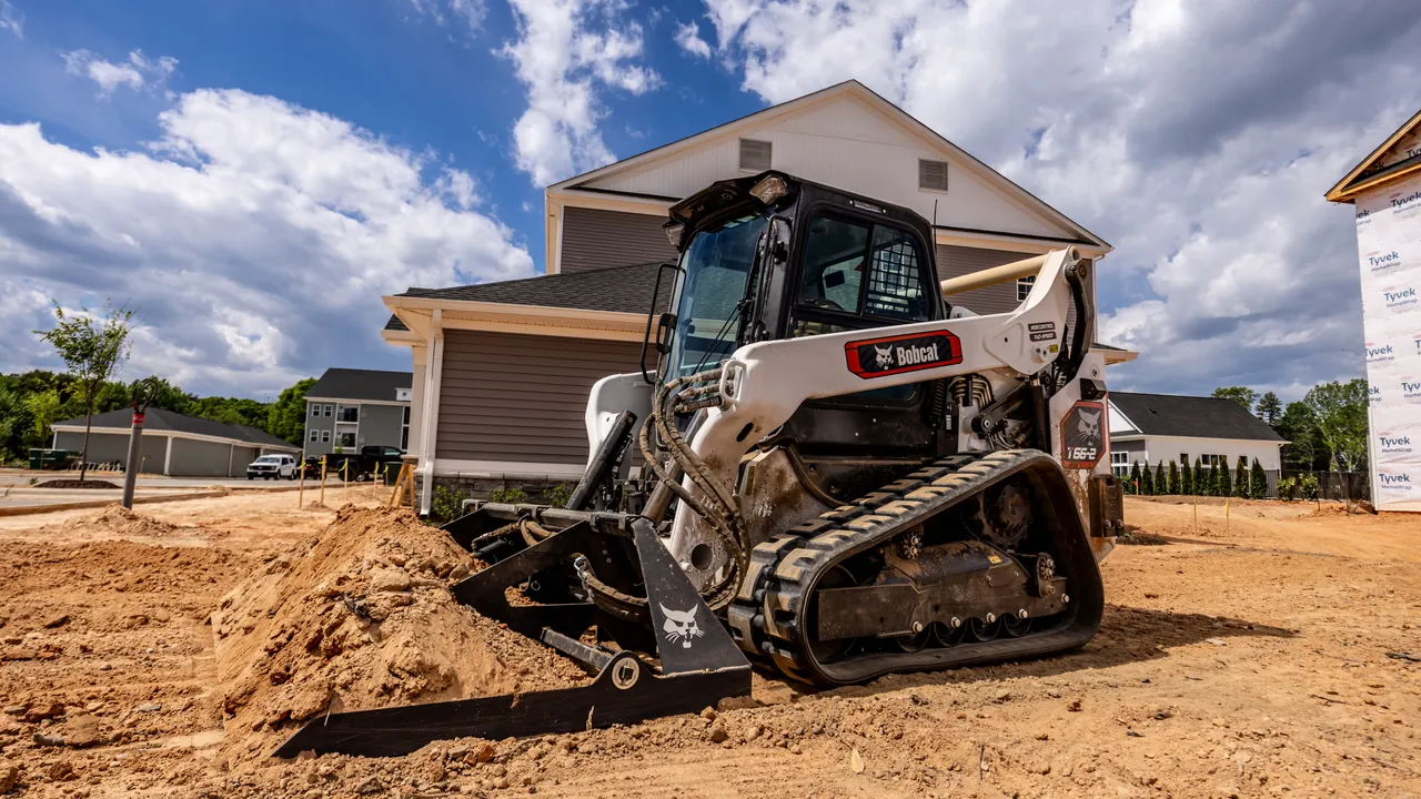 Tracked skid steer rough grading loose dirt on a cleared building site beside new houses