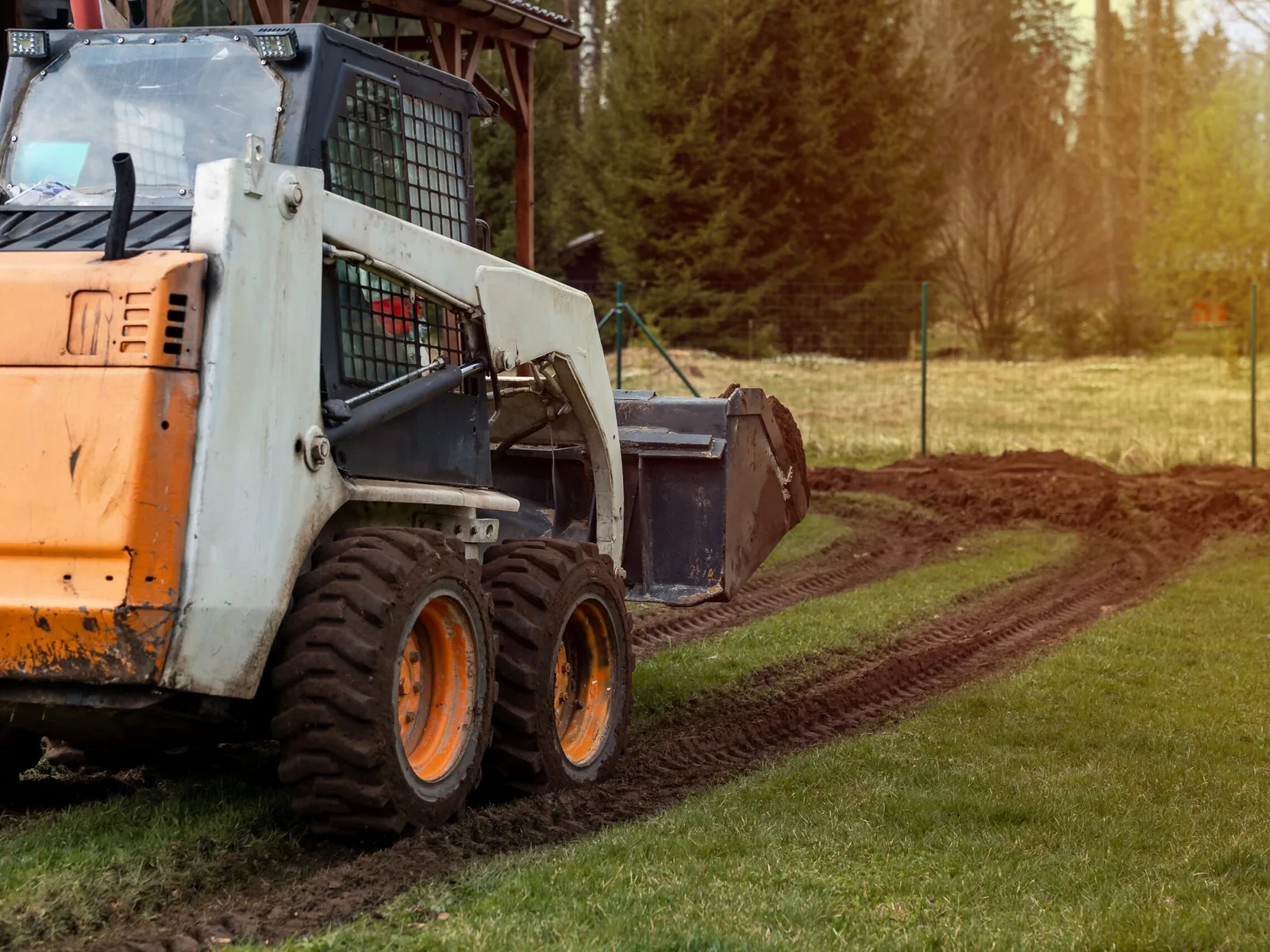 Wheeled skid steer with a loader bucket pushing loose soil across a grass field, leaving a low windrow of dirt and track ruts in front of a conifer treeline